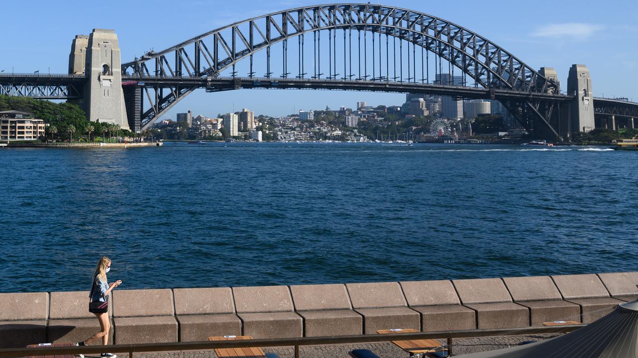 A lone woman wearing a protective face mask walks through a quiet Opera Bar in front of the Sydney Harbour Bridge in March 2020.